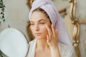 Woman with a head towel applying moisturiser at a mirror